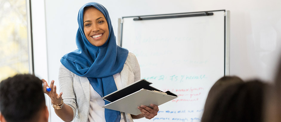 Lehrerin mit Kopftuch steht mit Tablet in der Hand vor einer Flipchart und unterrichtet