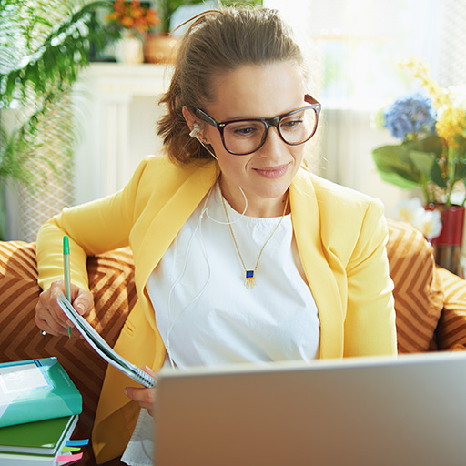 Eine Frau sitzt am Laptop mit Kopfhörer im Ohr und Stift in der Hand.