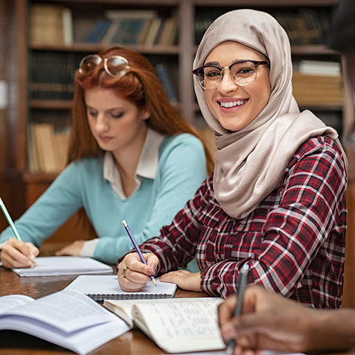 Frau mit Kopftuch sitzt in einer Bibliothek und lächelt in die Kamera