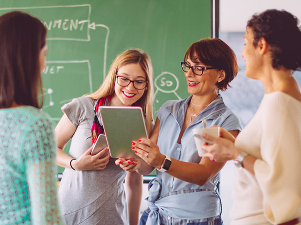 Mehrere Frauen stehen in einem Klassenraum und zeigen sich gegenseitig ihr Handy und Tablet-PC. 